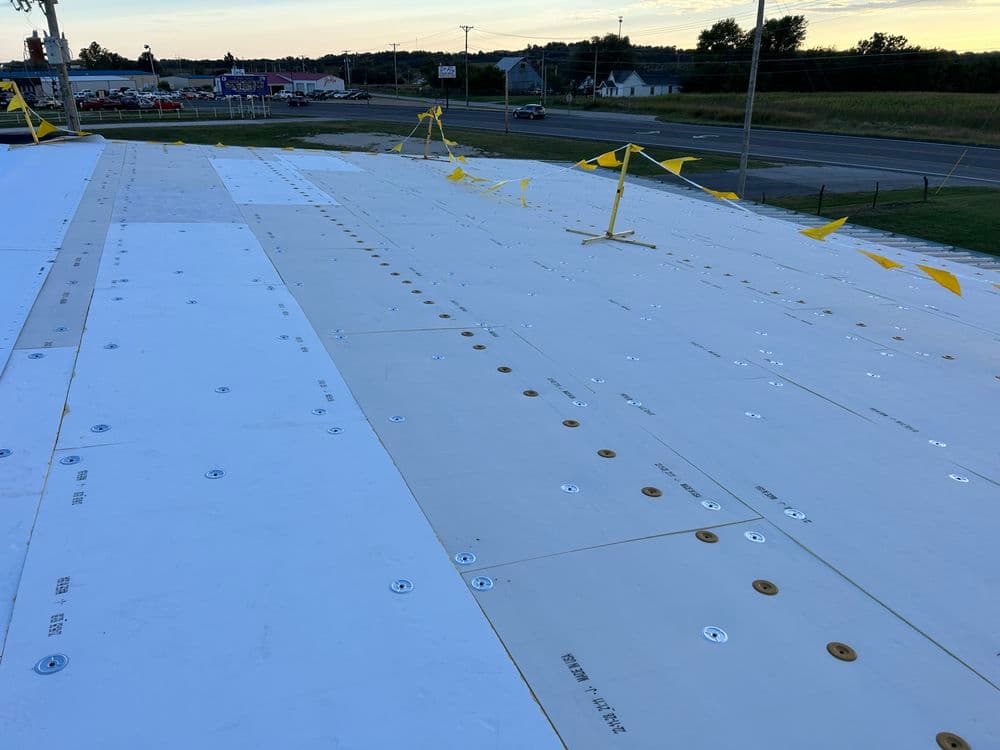 Roofing installation with white underlayment and safety barriers near a roadway.