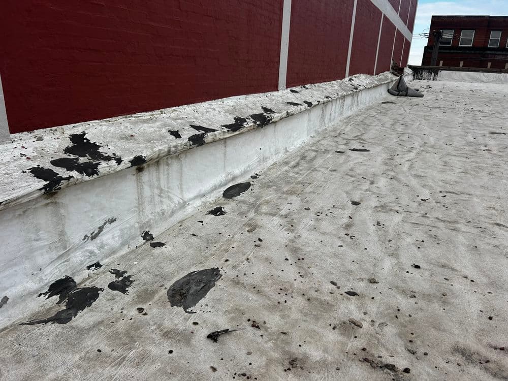 Roof surface with black streaks and patches against a red brick wall, showing wear and tear.