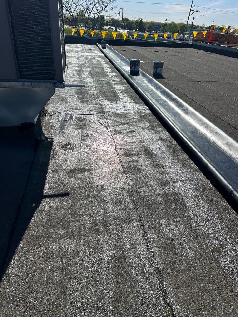 Roofing surface with shiny sealant; rolled materials and bright safety cones in background.