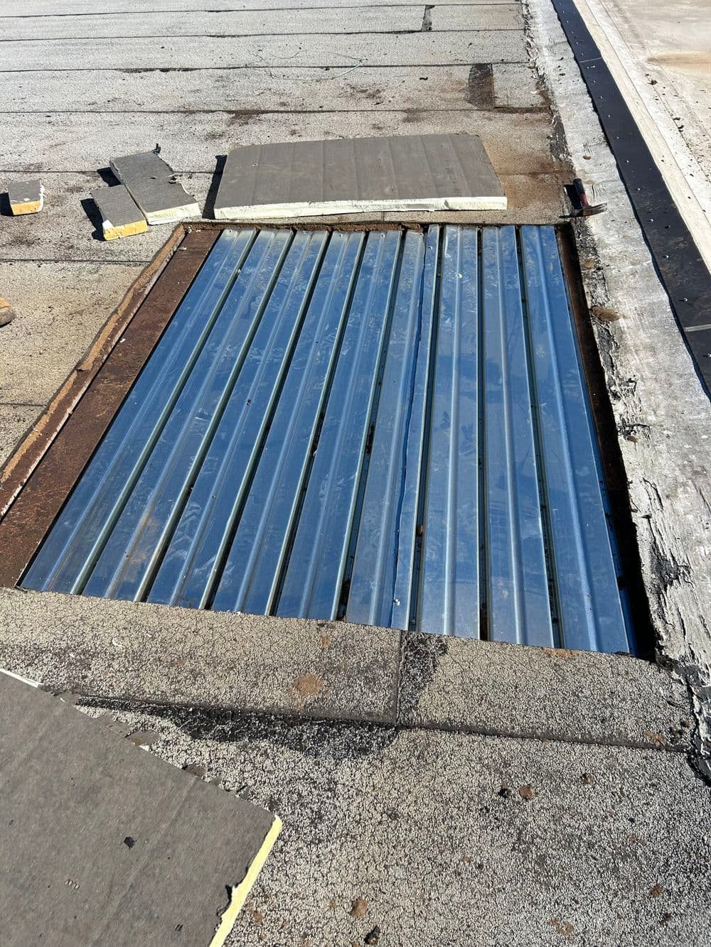 Open metal trench cover on a flat roof, revealing drainage and roofing materials below.
